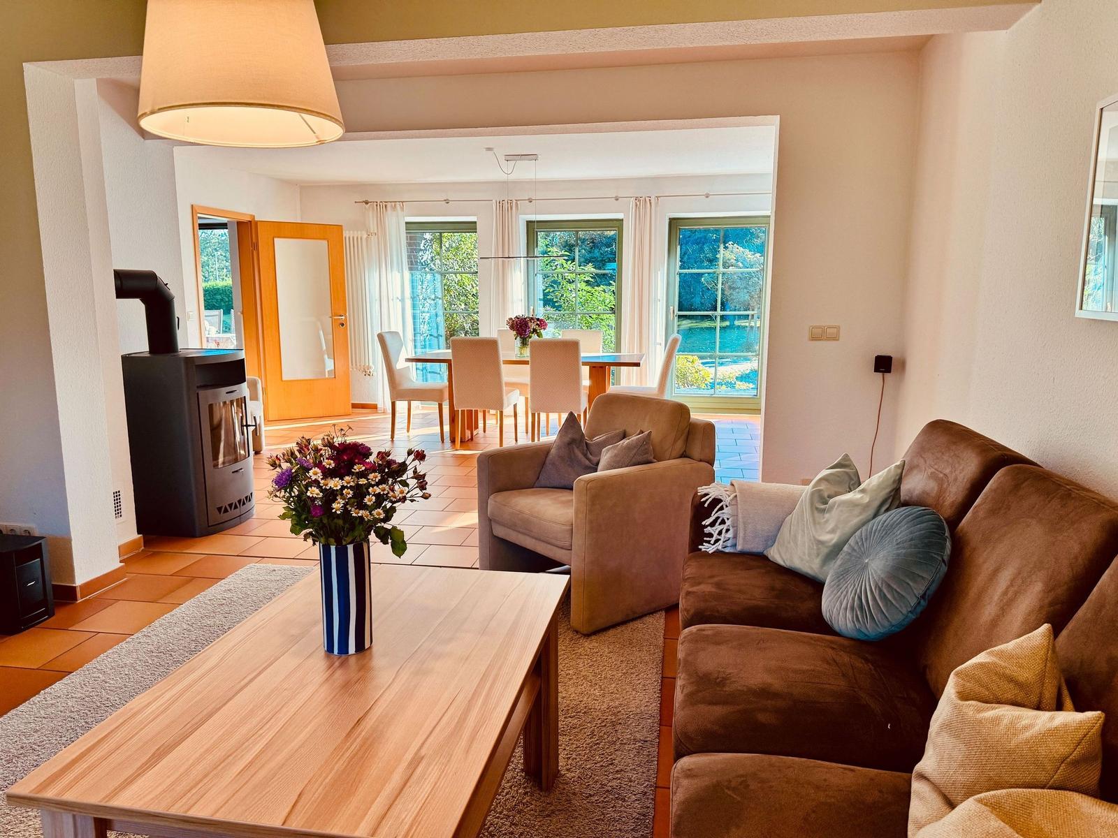 Wohnzimmer mit Sofa, Sessel und Holztisch. Im Hintergrund Essbereich mit Blick auf Garten.