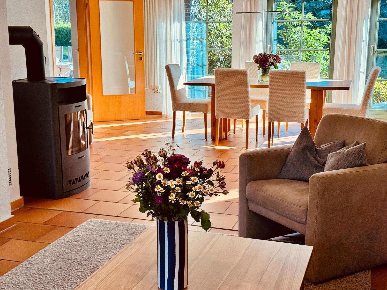 Wohn- und Essbereich mit Holztisch, Sofa und Kaminofen. Große Fenster mit Blick auf Garten.