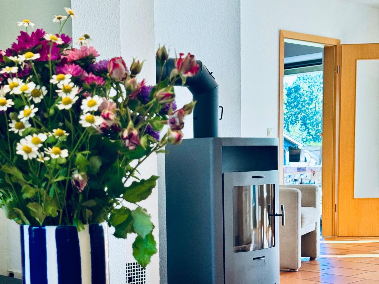 Kaminofen neben Blumenstrauß, Blick in Wohnbereich mit Sitzgelegenheit.