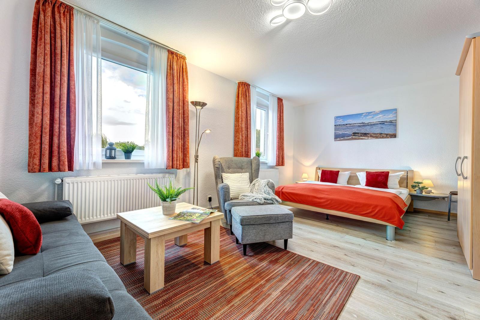 Schlafzimmer mit Bett, Sessel, Couch und Fenstern mit Vorhängen.