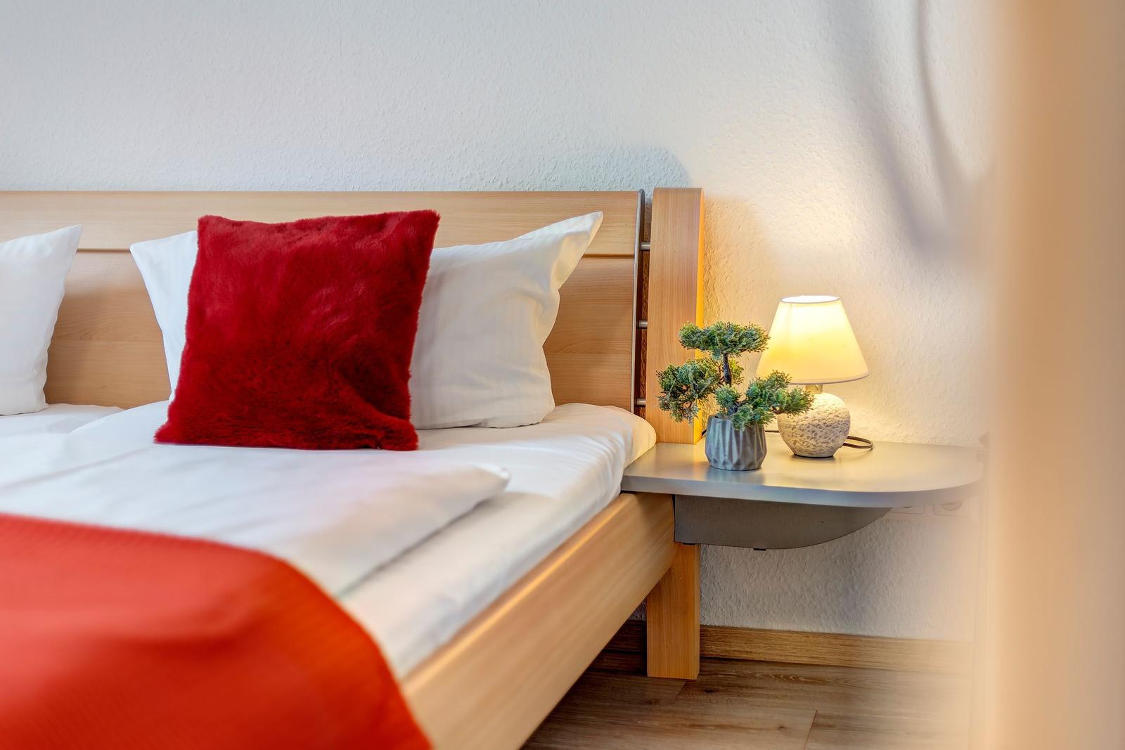 Hölzernes Bett mit weißen Kissen und rotem Kissen. Nachttisch mit Lampe und Pflanze.