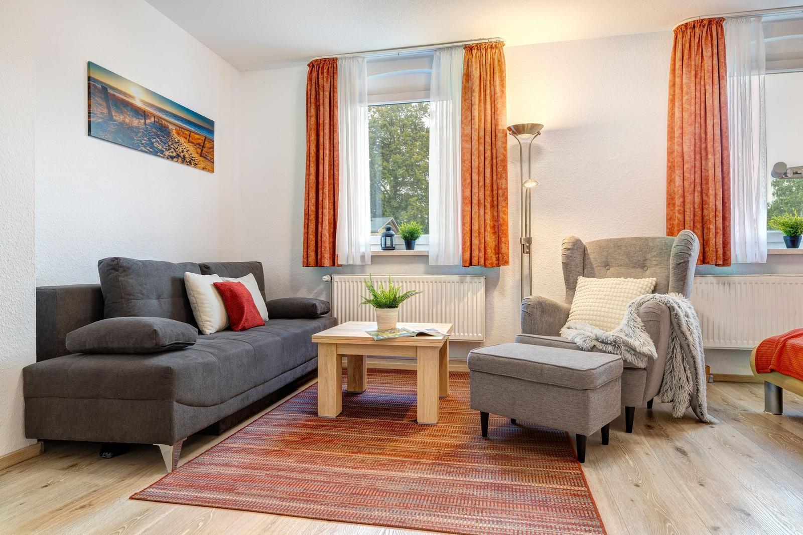 Wohnzimmer mit Sofa, Sessel und Holztisch. Fenster mit orangefarbenen Vorhängen.