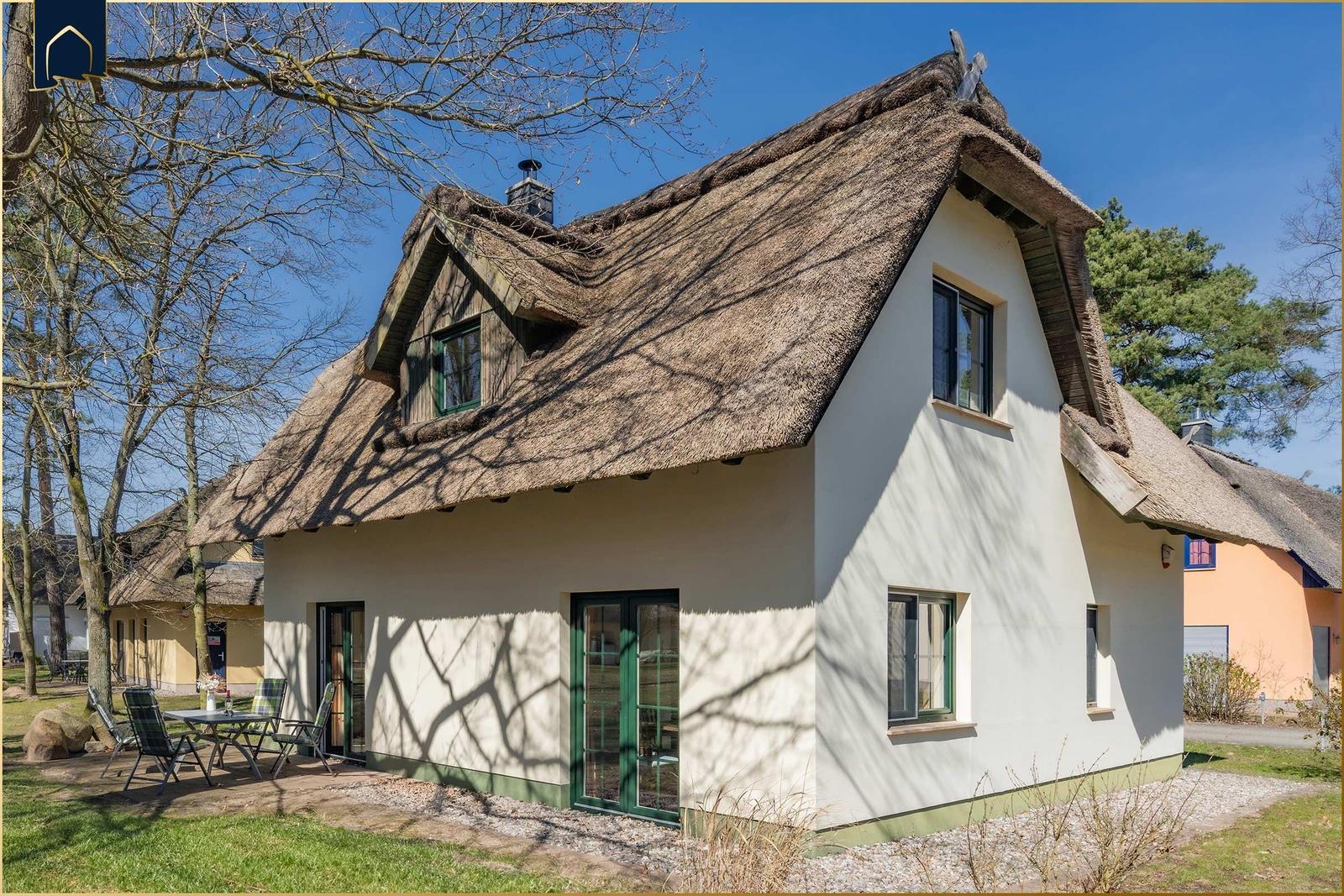 Haus mit Strohdach, grünen Fenstern und Terrasse mit Tisch und Stühlen.