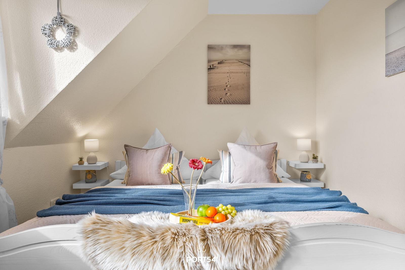 Schlafzimmer mit Dachschrägen, Bett mit Kissen und Decke, Wandbild und Nachttische mit Lampen.