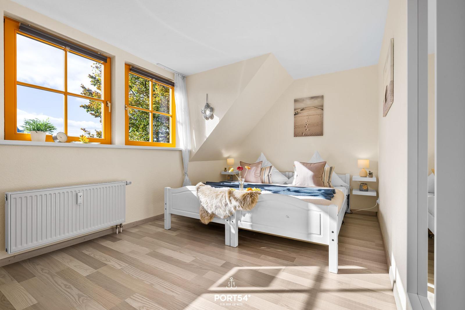 Schlafzimmer mit weißem Bett, Holzboden und zwei Fenstern mit orangefarbenen Rahmen.