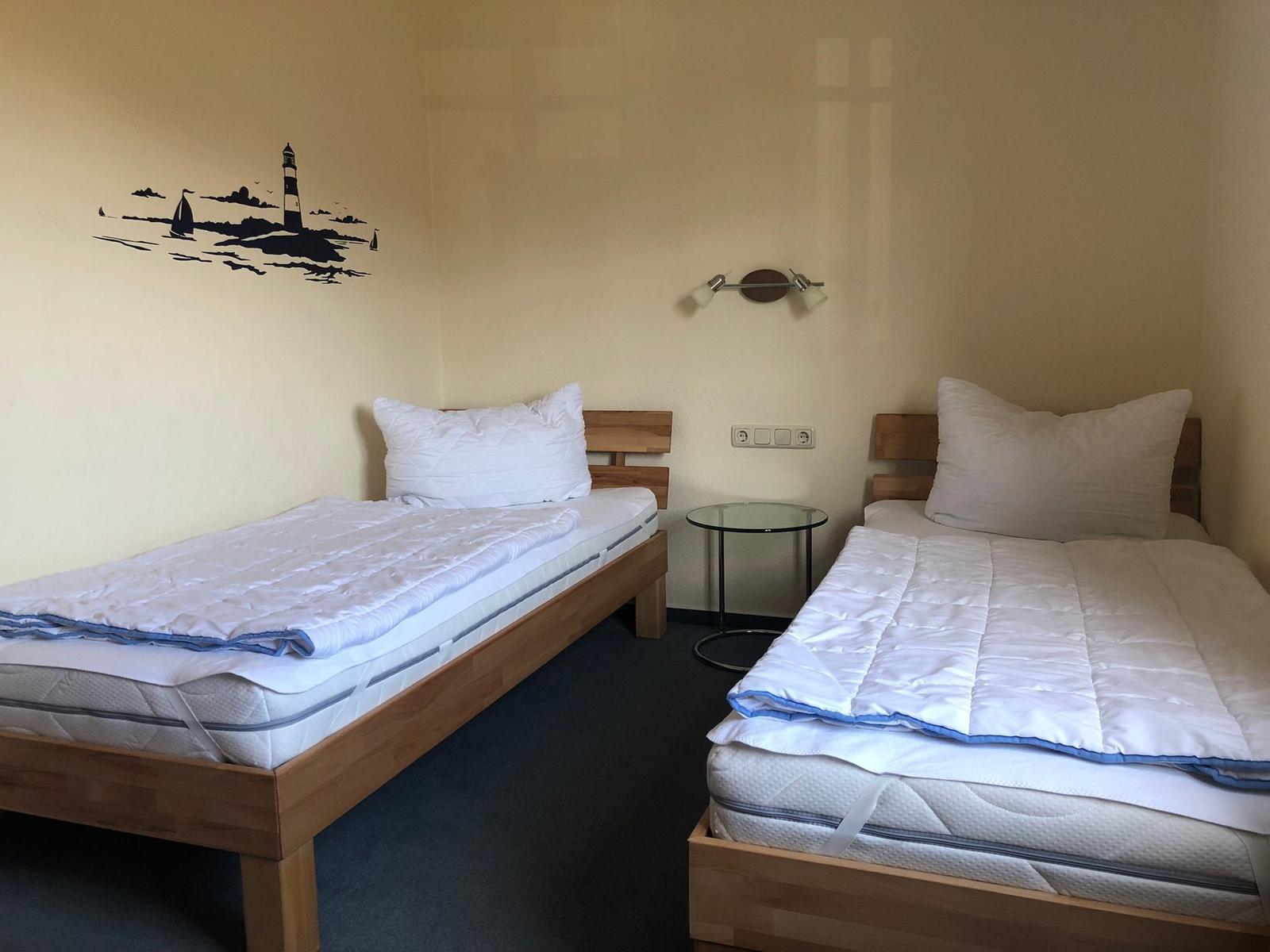 Zwei Einzelbetten mit Matratzen und Kissen. Wanddekoration mit Leuchtturm. Zwischen den Betten steht ein kleiner Tisch.
