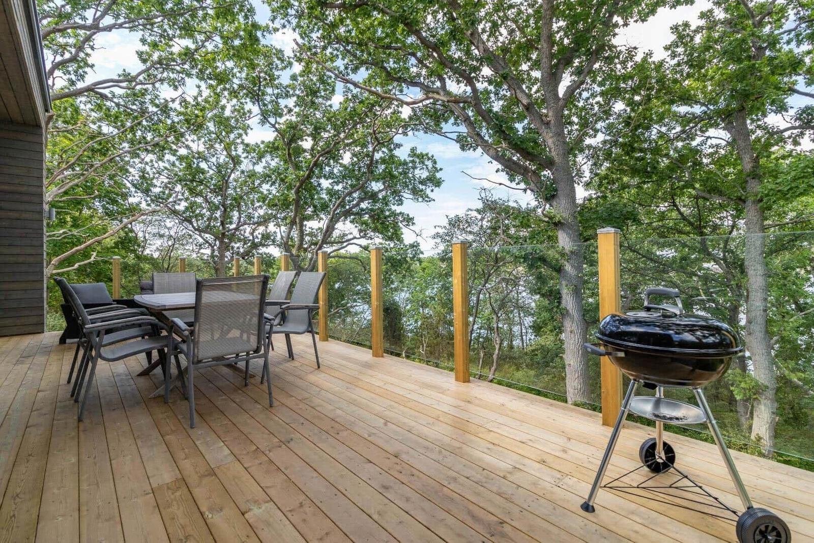 Holzterrasse mit Grill, Tisch und Stühlen unter Bäumen.