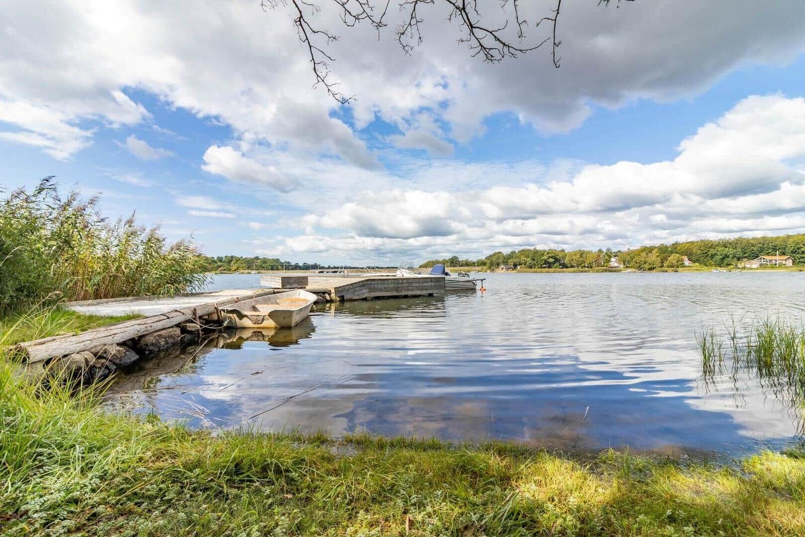 Boot liegt am Steg eines ruhigen Sees mit Gras und Bäumen im Hintergrund.