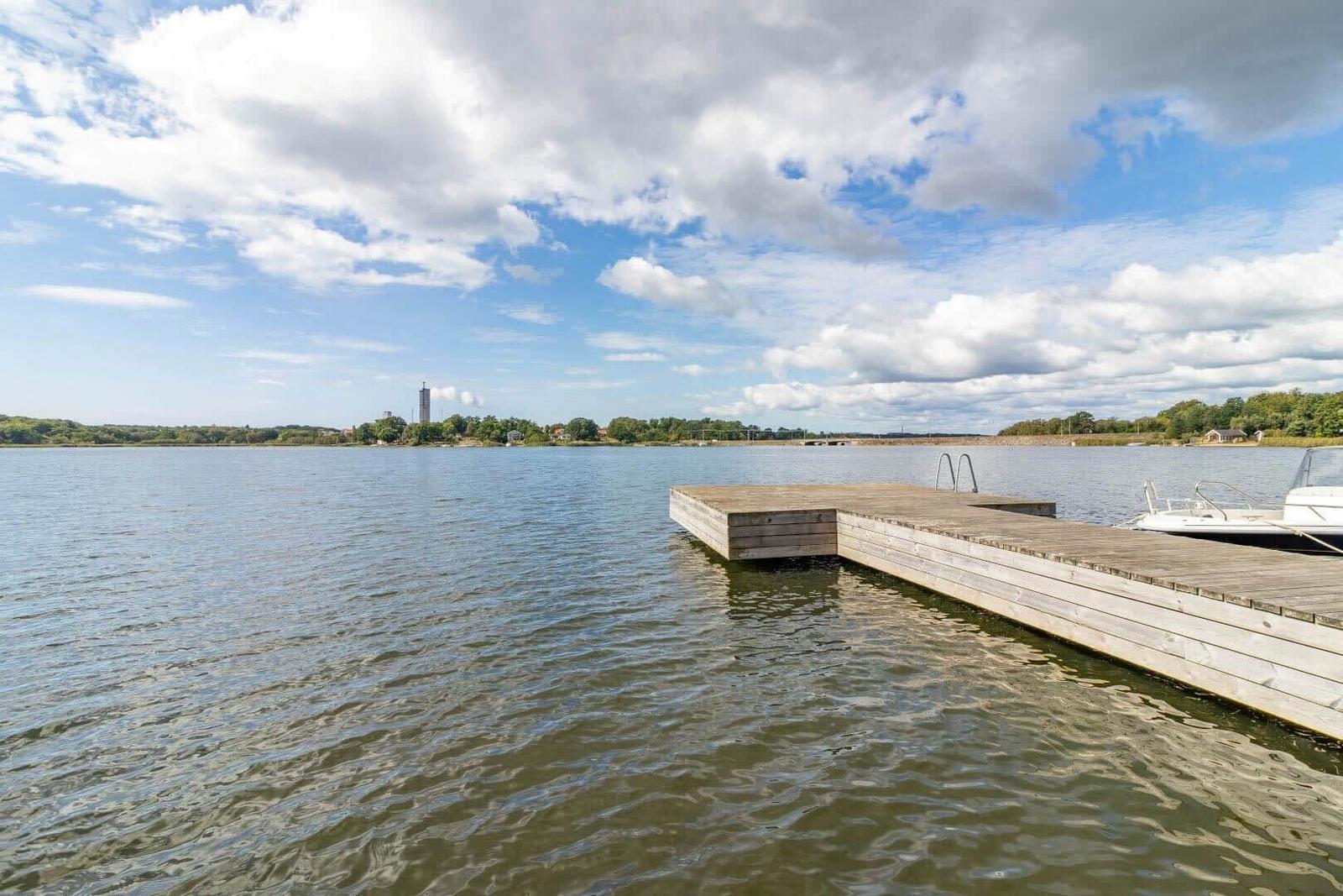 Holzsteg mit Boot liegt am Wasser. Im Hintergrund sind Bäume und ein Turm sichtbar.
