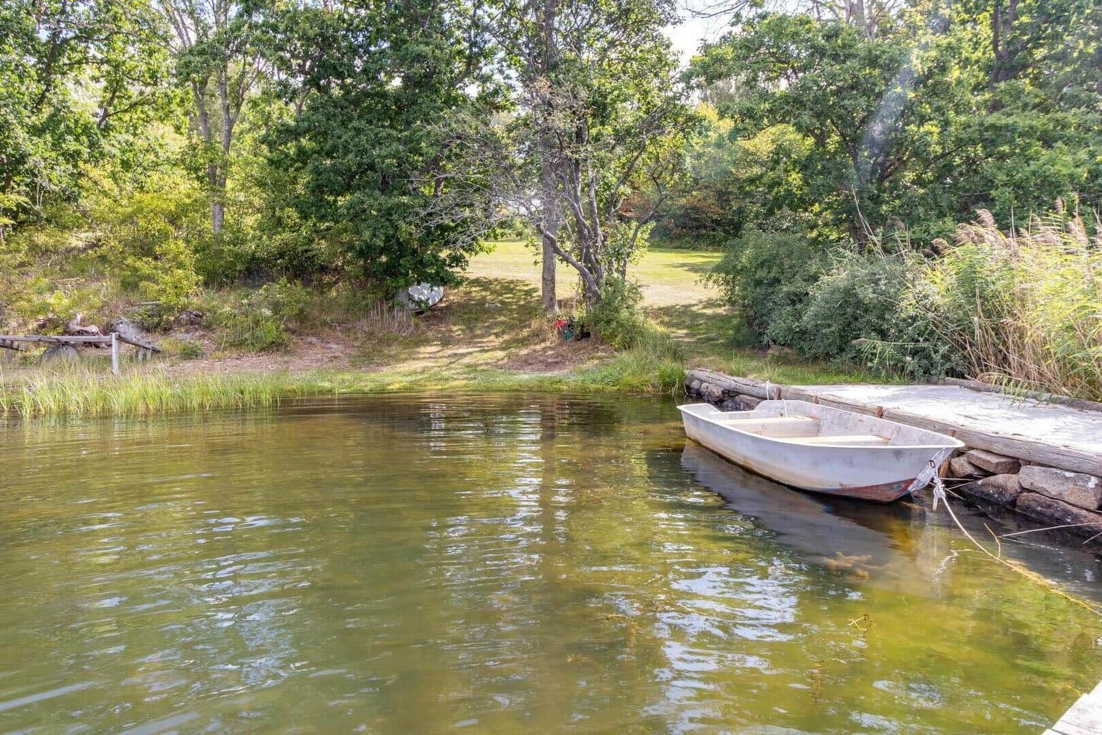 Ein kleines Boot liegt an einem Holzsteg am Ufer eines ruhigen Gewässers, umgeben von Grünflächen und Bäumen.