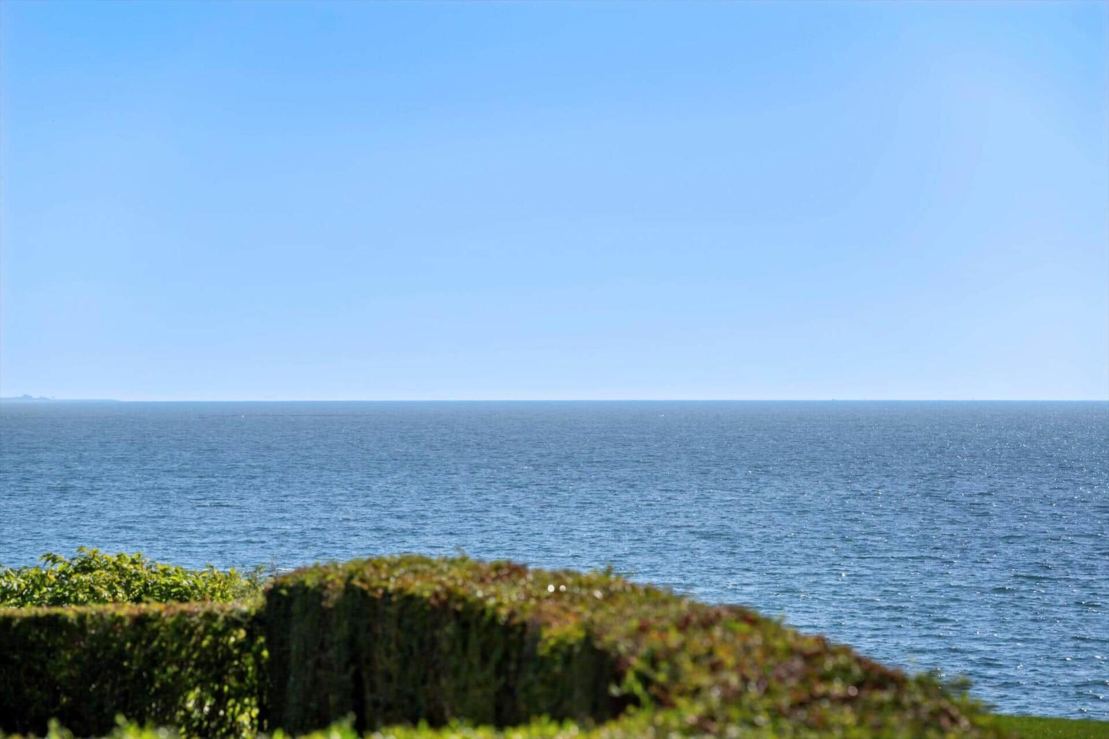 Weitläufiger Blick auf das Meer unter blauem Himmel mit grüner Hecke im Vordergrund.