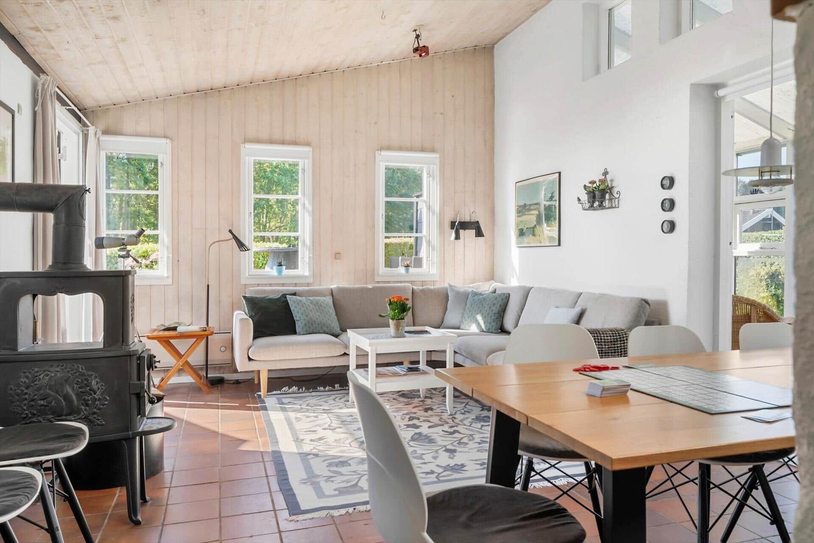 Wohn- und Essbereich mit Sofa, Esstisch und Kaminofen. Holzwände und Fenster mit Blick auf Grün.