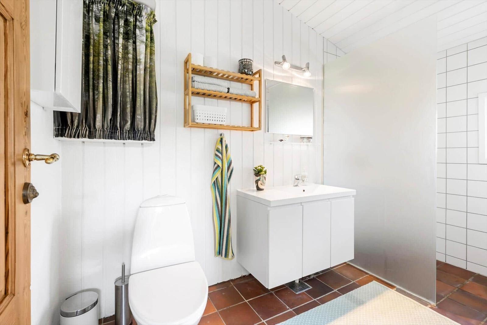 Badezimmer mit Toilette, Waschbecken und Dusche. Weiße Wände, brauner Boden und Holzregal.