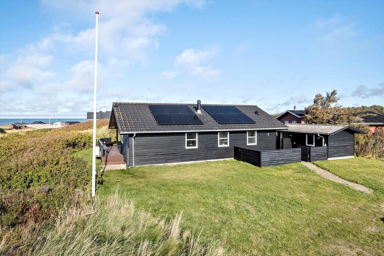 Schwarzes Holzhaus mit Solarpanelen, Grünfläche und Blick aufs Meer.