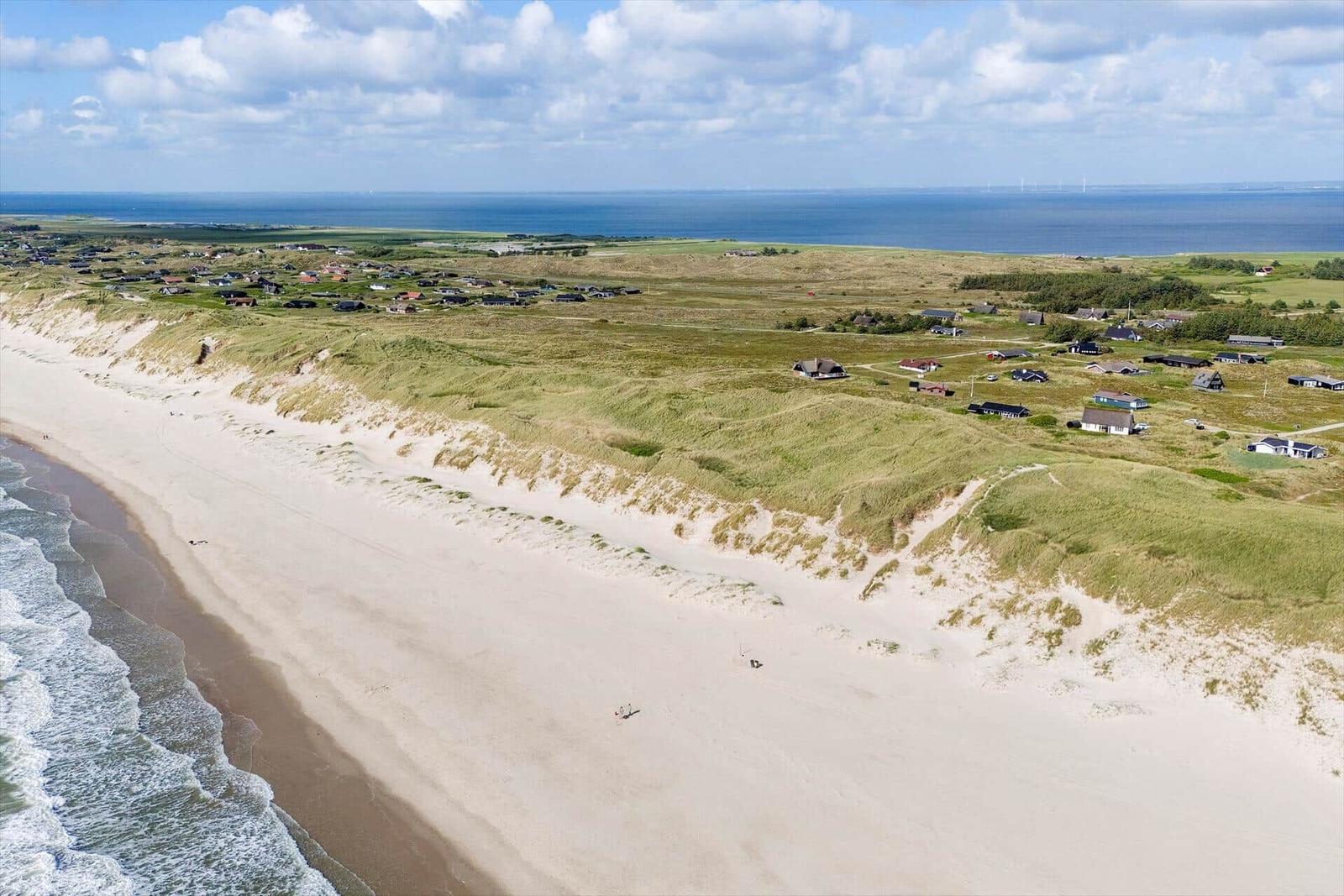 Strand mit Dünen und Häusern am Meer unter blauem Himmel.