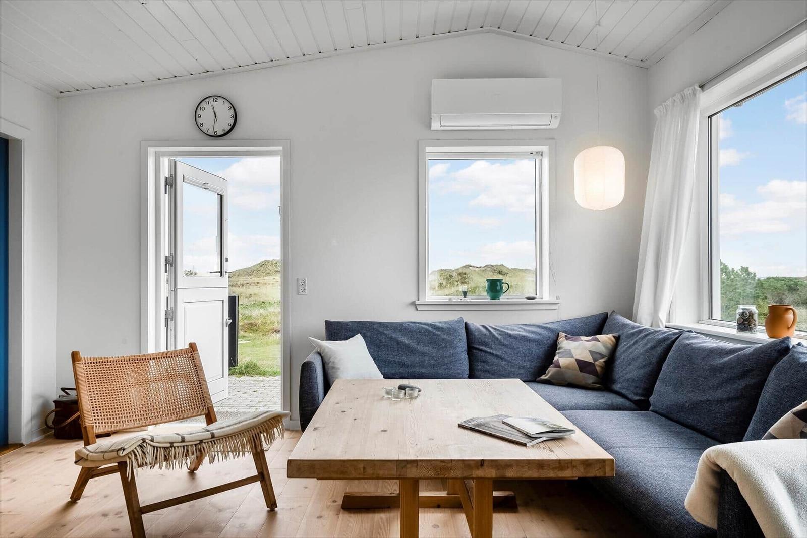 Wohnzimmer mit Sofa, Holztisch und Sitzgelegenheit. Fenster und Tür zeigen Landschaft.