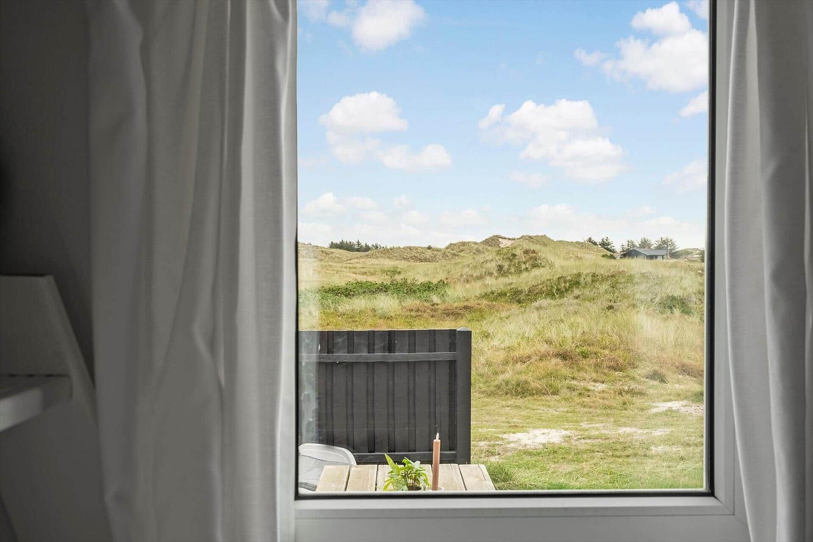 Durch das Fenster blickt man auf eine grüne Dünenlandschaft unter blauem Himmel.