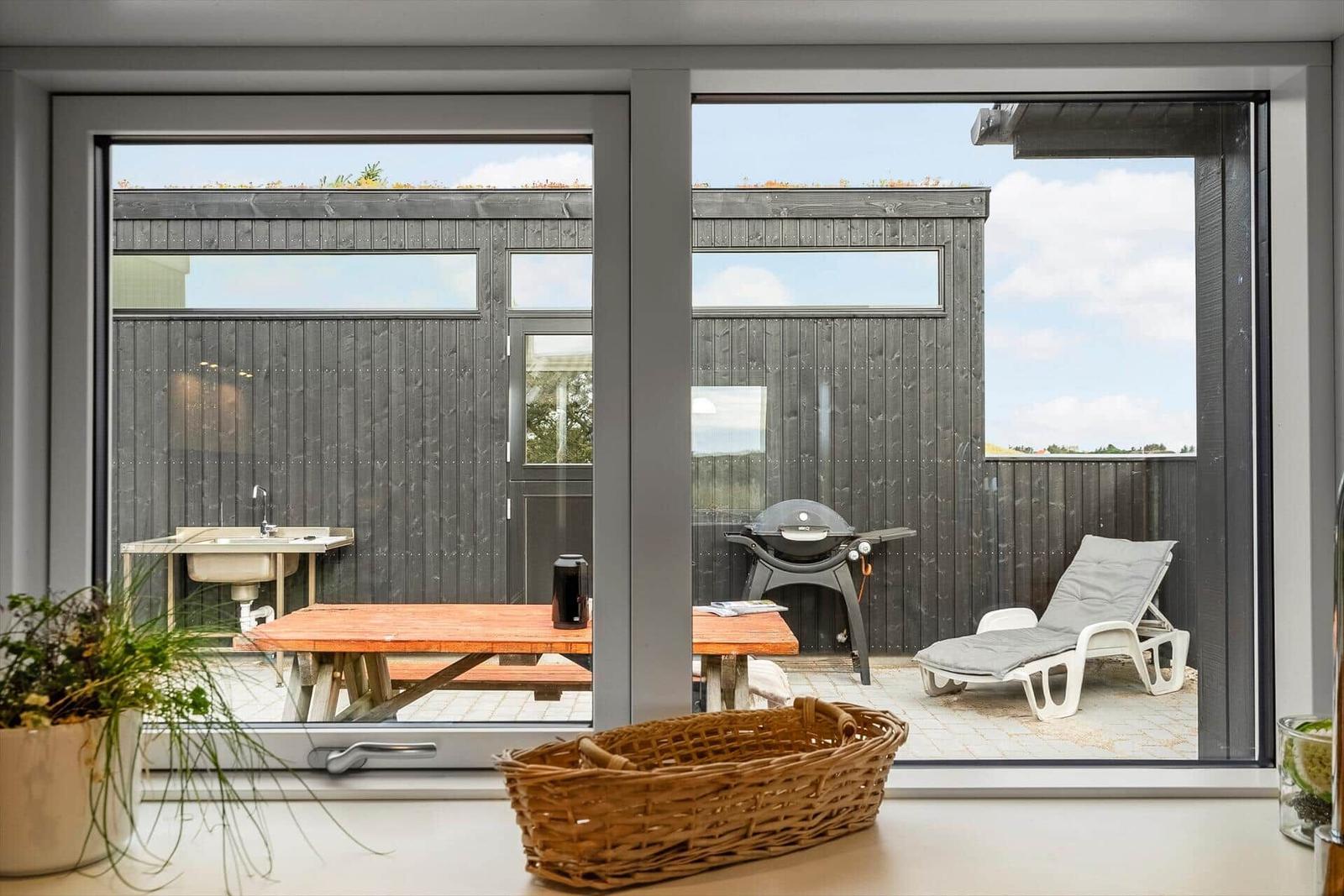 Terrasse mit Grill, Liegestuhl und Holztisch. Blick auf dunkle Holzwände und Himmel.