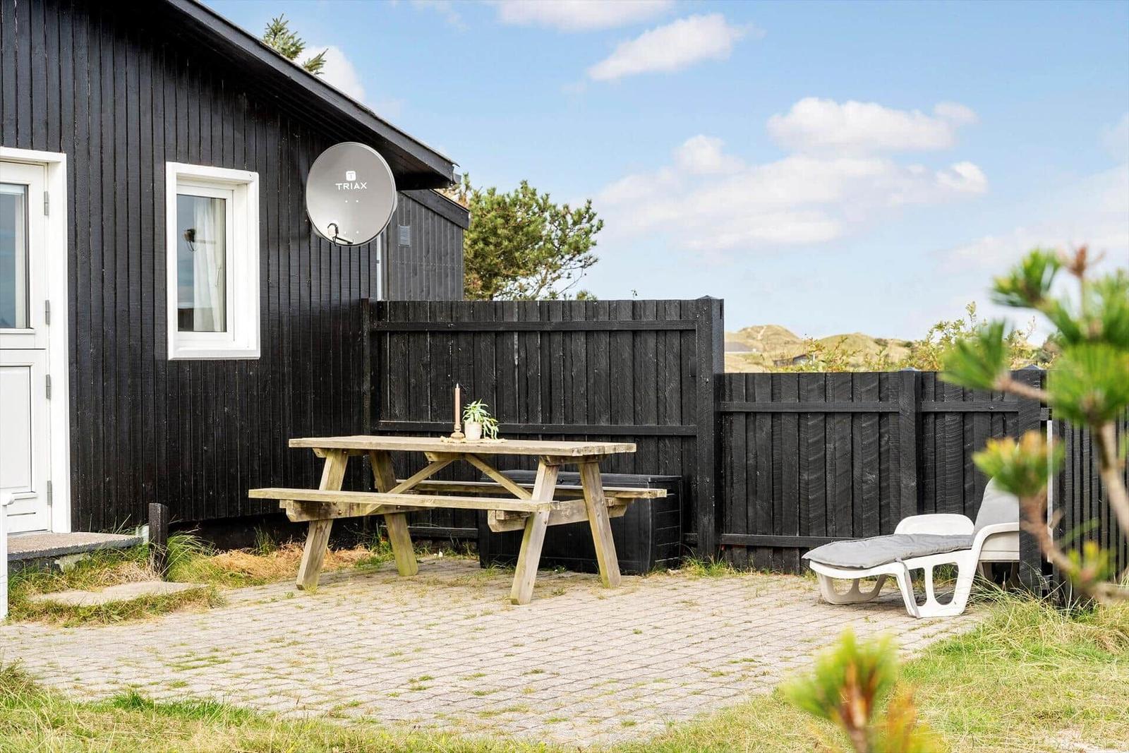 Außenbereich mit Tisch, Liegestuhl und Holzhaus. Hinterhof mit Zaun und Strandblick.
