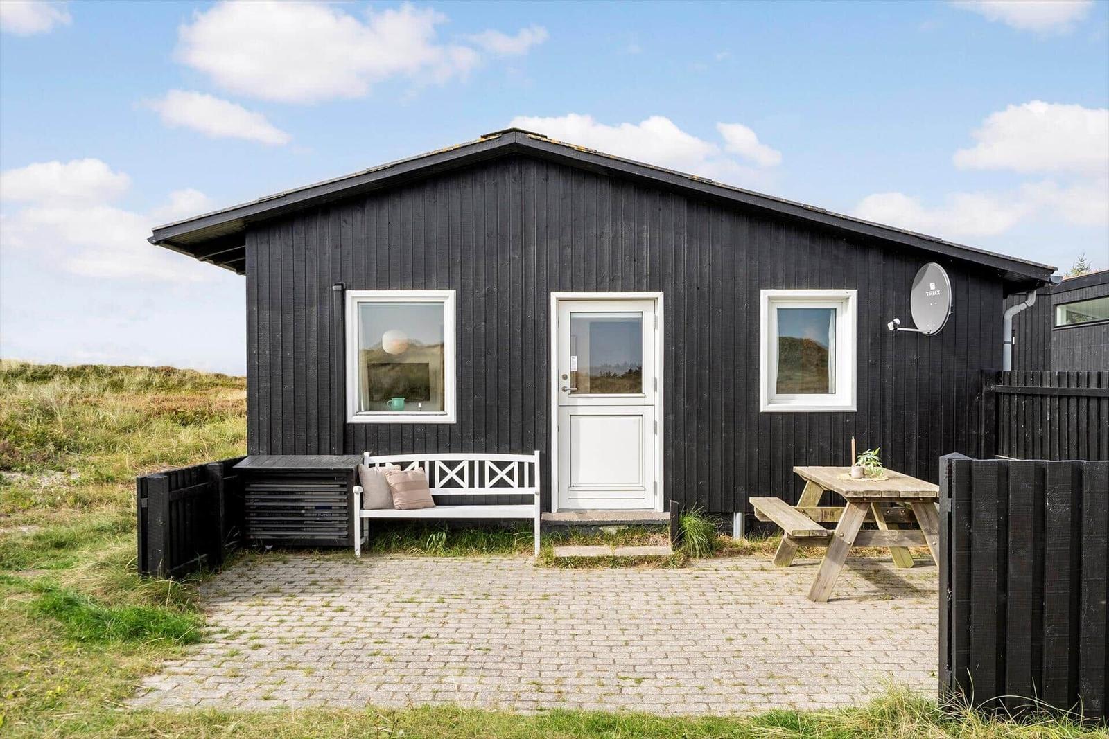 Schwarzes Holzhaus mit weißer Tür und Fenstern. Außenbereich mit Bänken und Tisch.