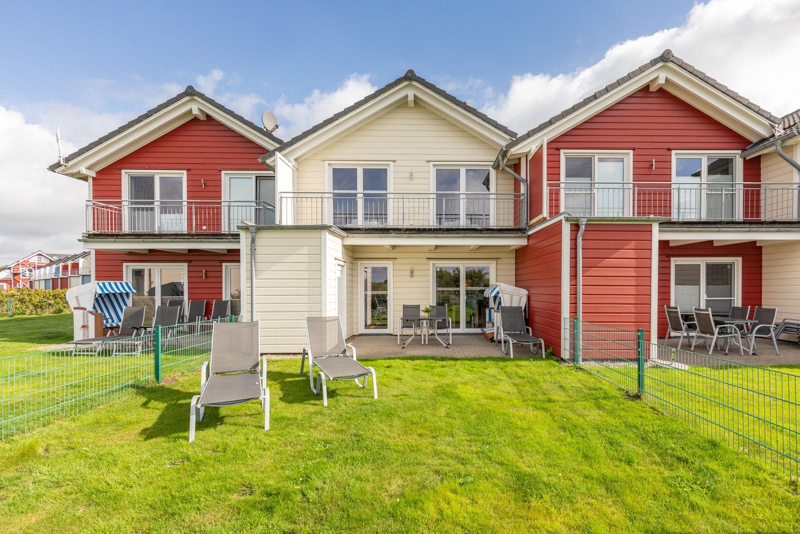 Red and cream-colored vacation home with balcony and garden area with lounge chairs.