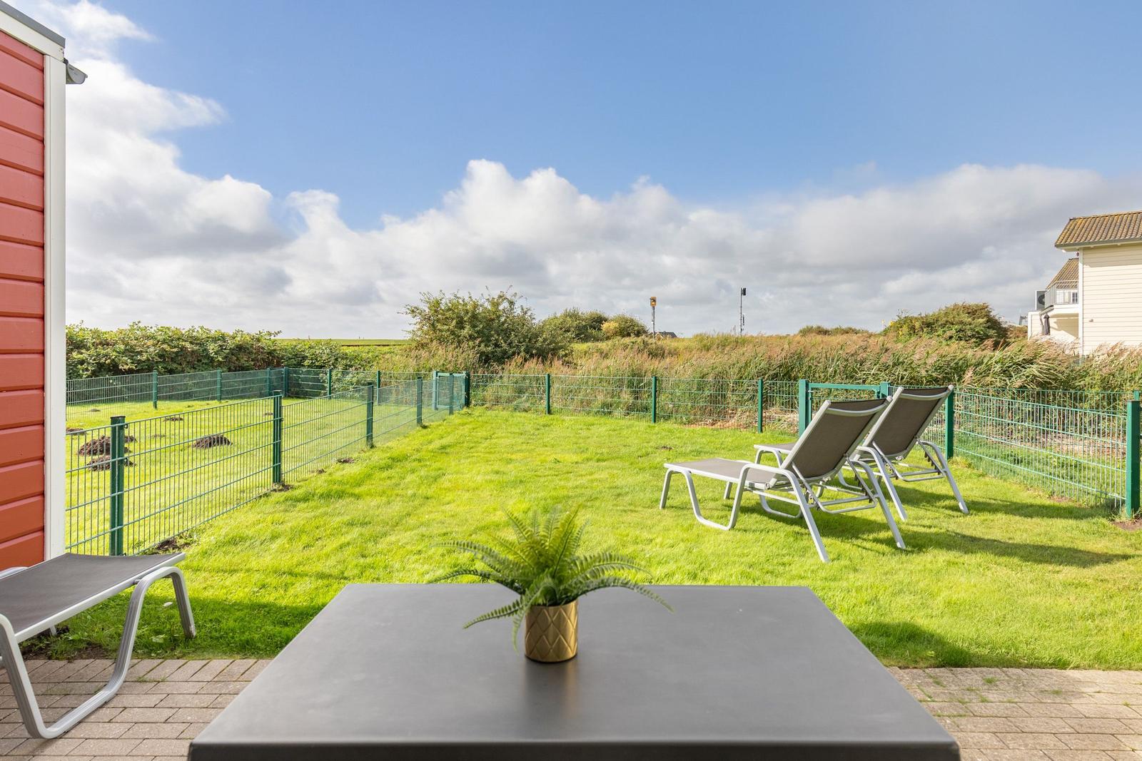 Garden with loungers and table. Green area behind fence. Sky with clouds.