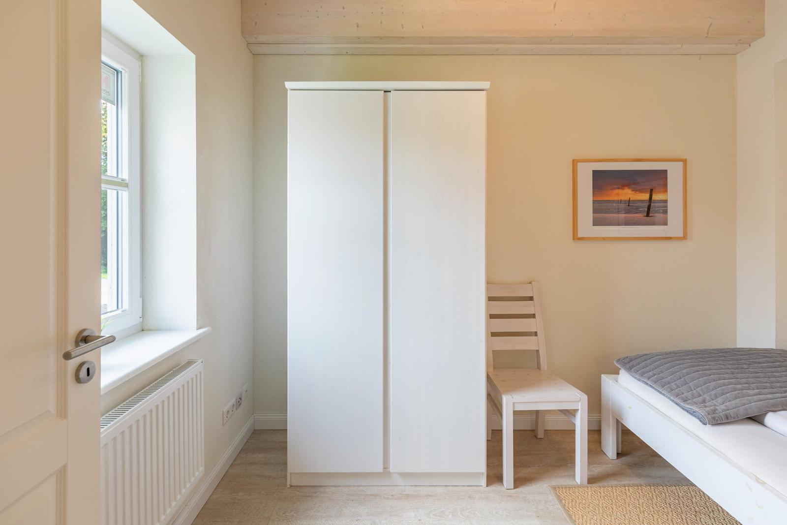 Bedroom with white wardrobe, chair, and bed. Window and wall picture are visible.