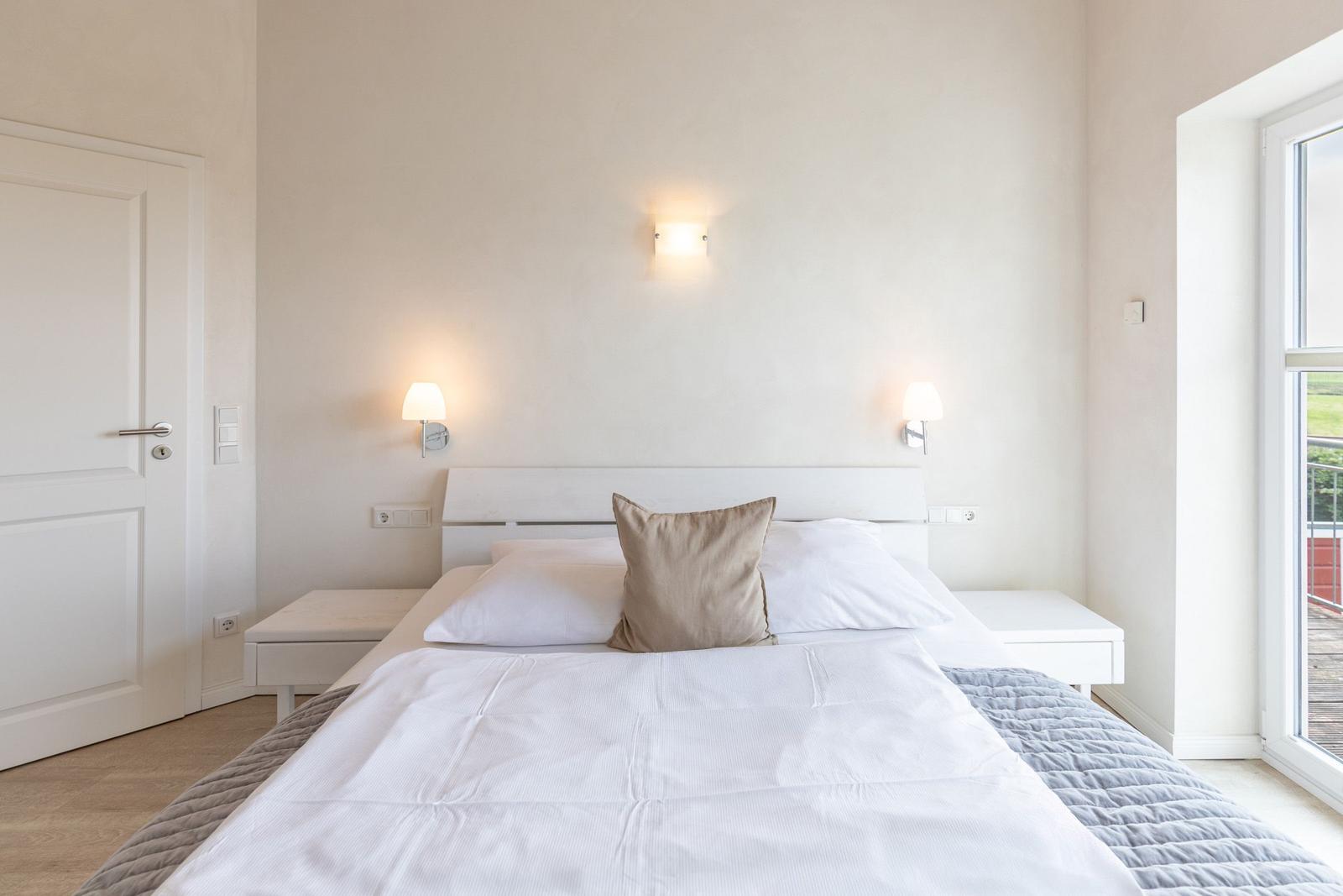 Bedroom with white bed, nightstands, and wall lamps. View to balcony through window.
