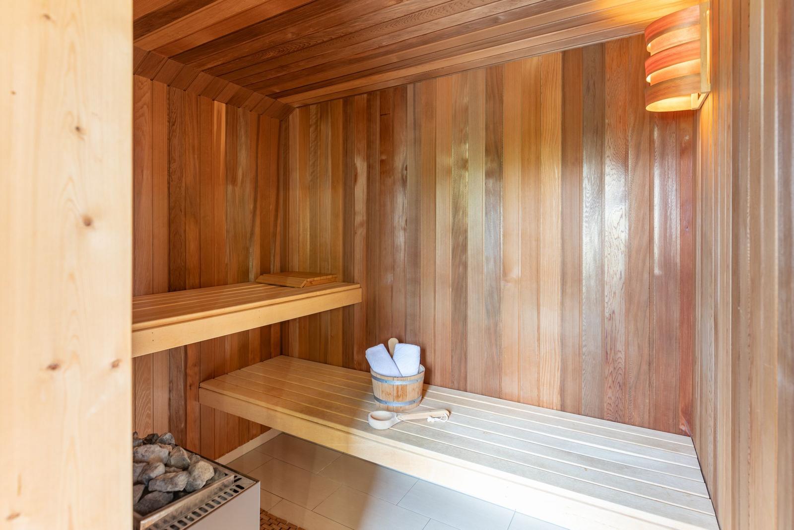 Sauna with wooden bench, bucket with towels, and wall lighting.