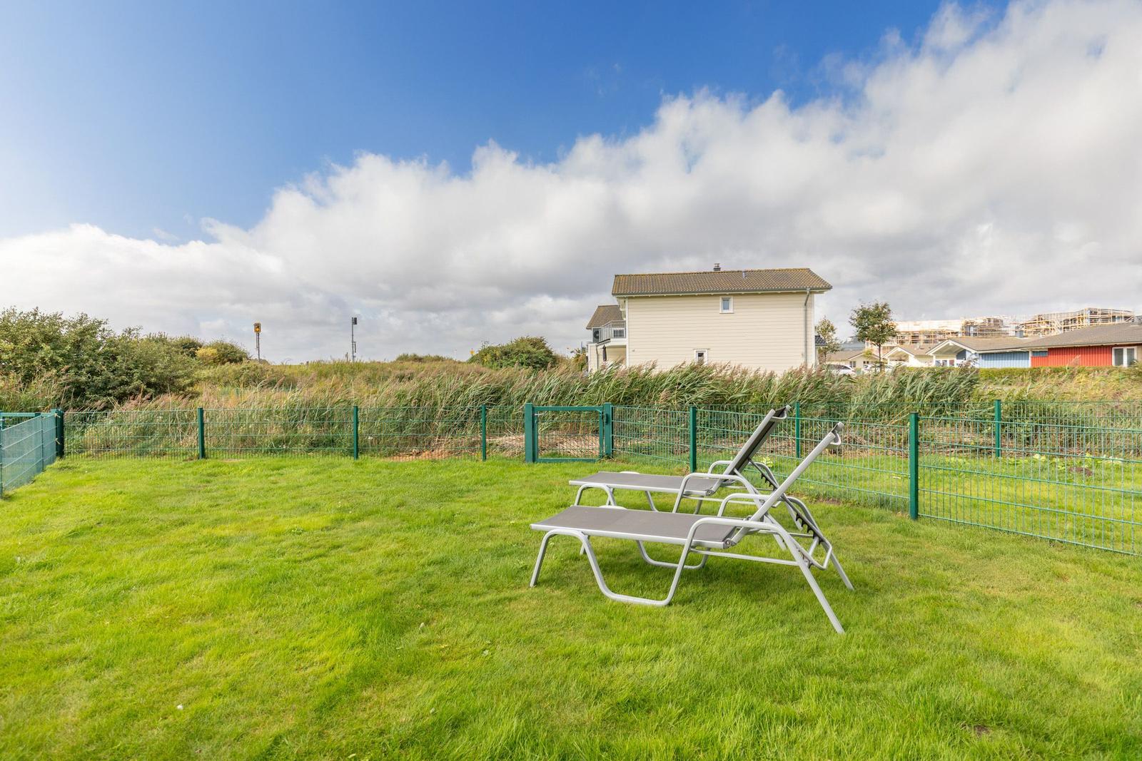 Green garden with sun loungers and fence. House and coastal vegetation in background.