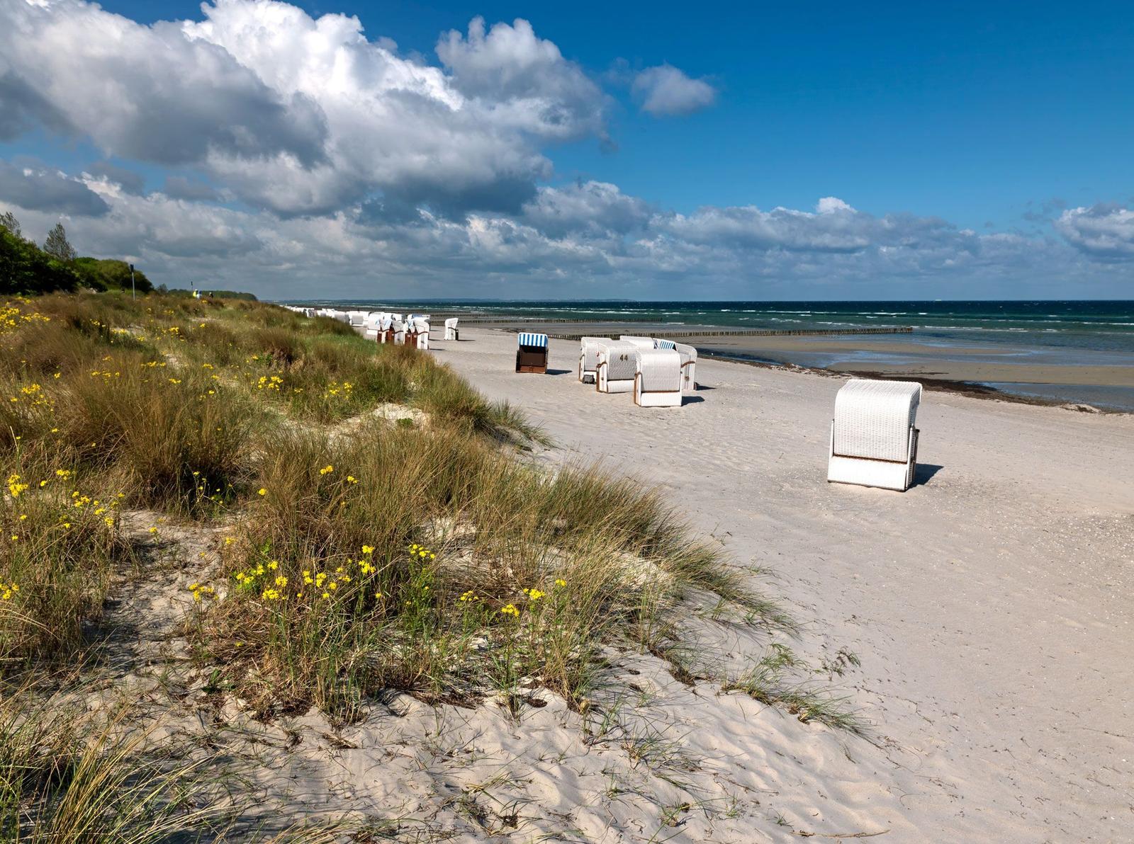 Strand mit Strandkörben, Dünen und Meer unter blauem Himmel mit Wolken.