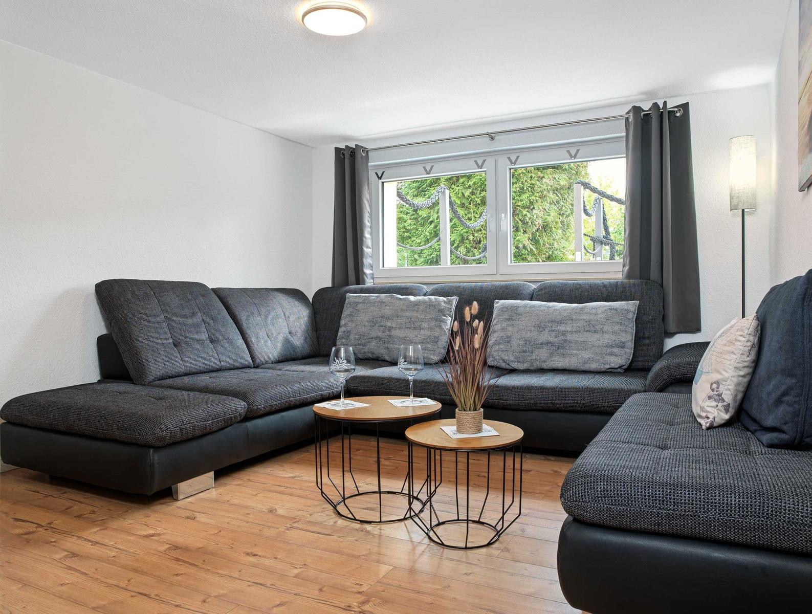 Ein Wohnzimmer mit grauem Sofa, Holzboden und Fenster mit Blick auf Grün.