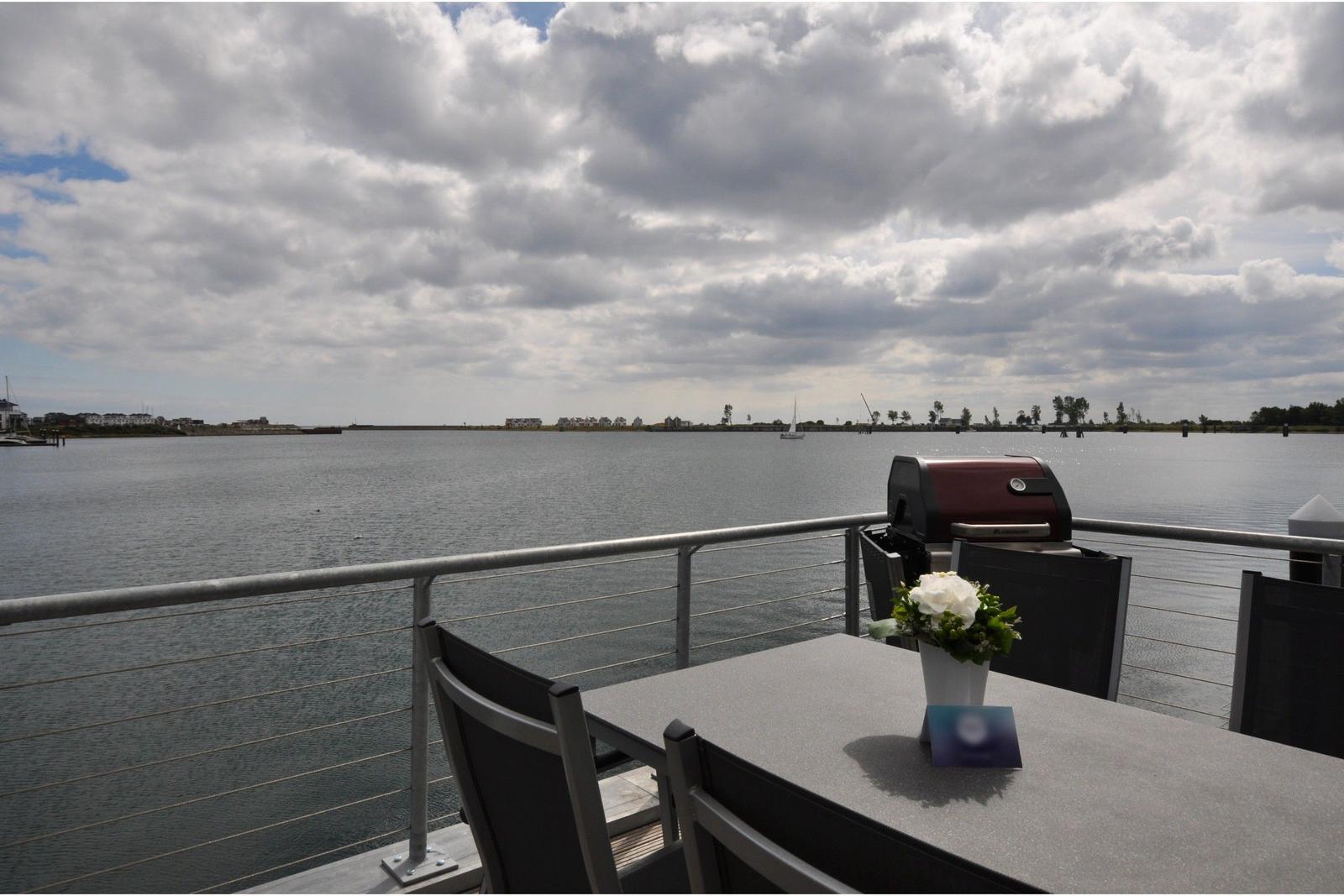 Terrasse mit Grill und Tisch am Wasser mit Blick auf See und Segelboot.