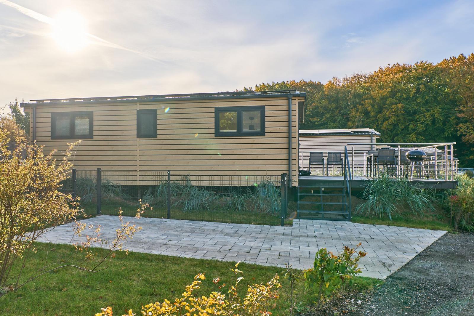 Moderne Holzhütte mit Terrasse, Grill und Gartenbereich. Sonniger Tag mit Wald im Hintergrund.