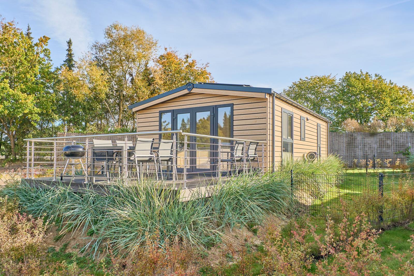 Holzhaus mit Terrasse, Grill und Garten. Umgeben von Bäumen und Grünflächen.
