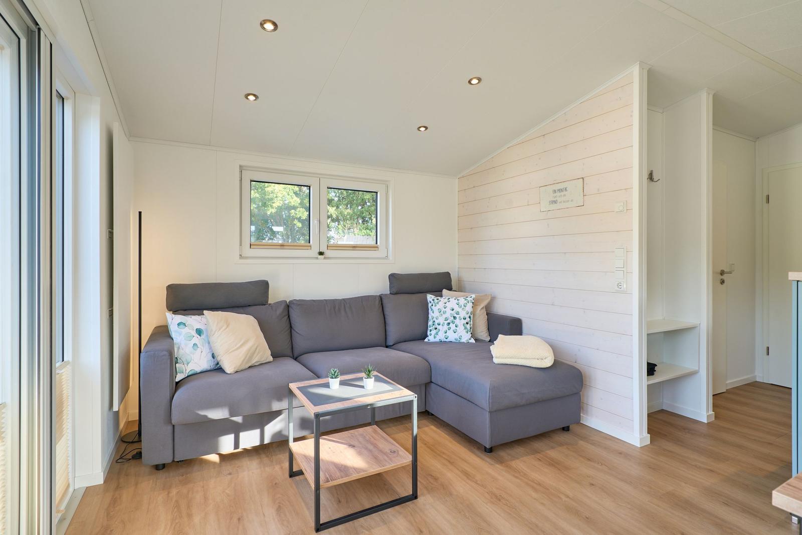 Ein Wohnzimmer mit grauem Sofa, Tisch und Fenster. Holzfußboden und Holzwand.