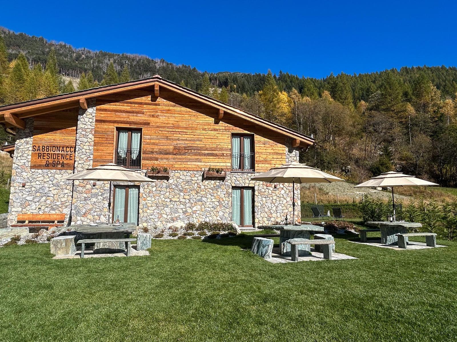 Haus mit Stein- und Holzfassade, Garten mit Tischen und Sonnenschirmen, Berglandschaft im Hintergrund.