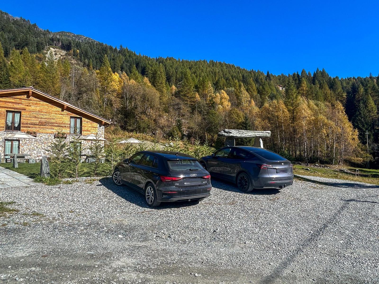 Zwei Autos parken vor einem Holzhaus in einem Wald mit Herbstfarben.