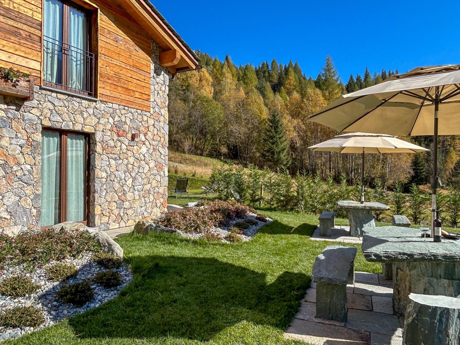 Haus mit Steinwand und Holzfassade, Terrasse mit Sonnenschirmen und Garten.