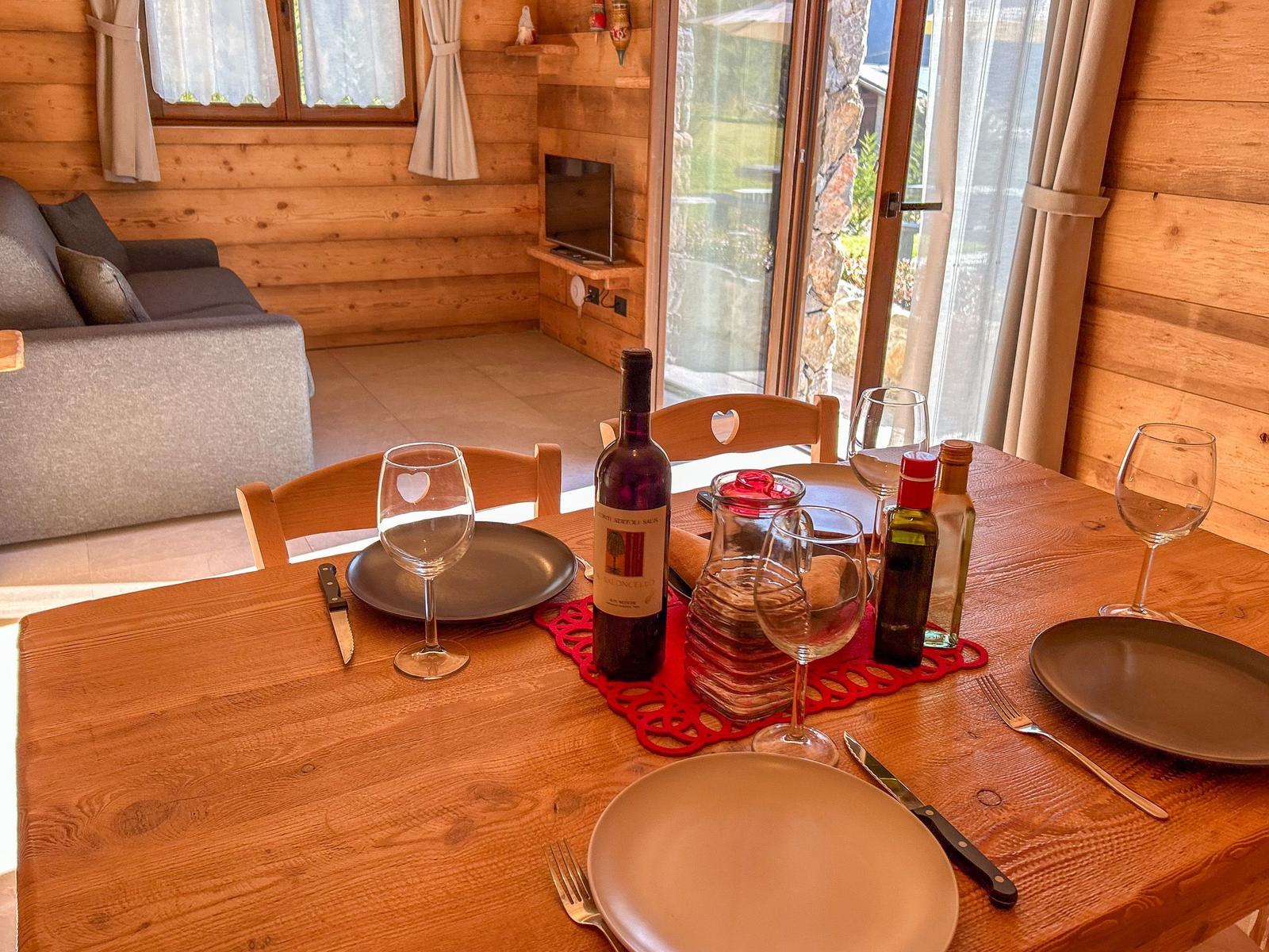 Holztisch mit Geschirr und Flaschen im Essbereich. Hintergrund: Wohnzimmer mit Sofa und TV.