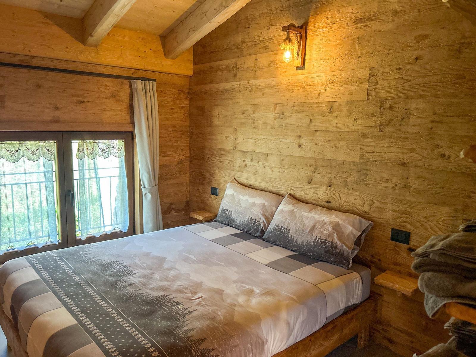 Schlafzimmer mit Holzwänden, Bett mit Bergmotiv und Fenster mit Blick auf Wald.
