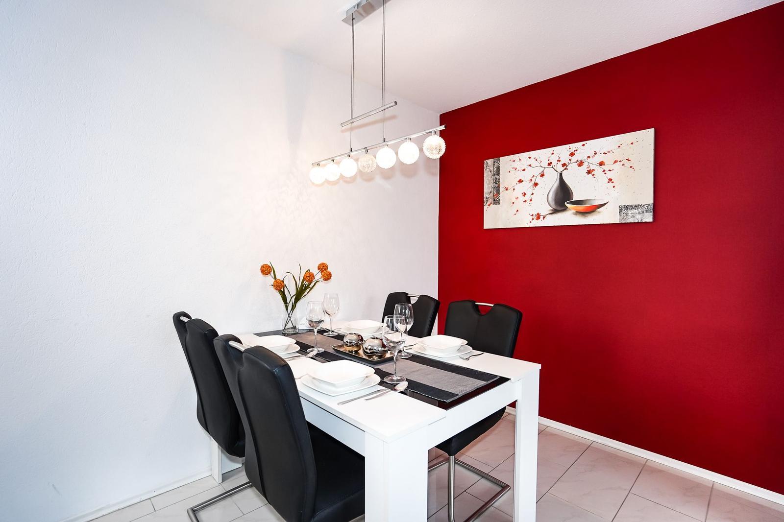 Esszimmer mit weißem Tisch, schwarzen Stühlen und roter Wand. Deckenleuchte und Wandbild.