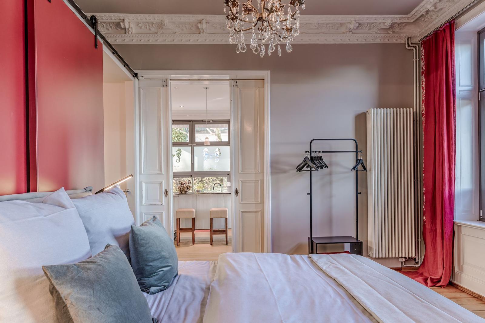 Schlafzimmer mit Bett, roten Vorhängen und Kronleuchter. Tür führt in Wohnbereich.