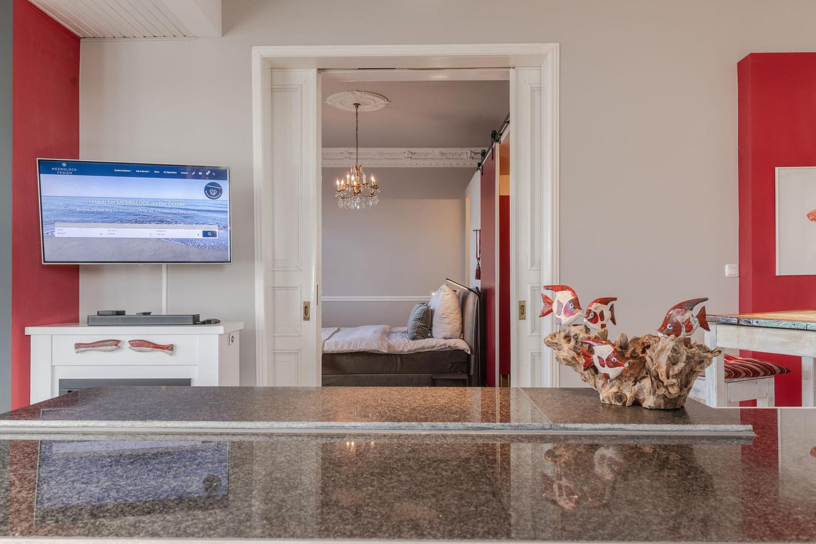 Wohnbereich mit Fernseher und Blick in Schlafzimmer mit Kronleuchter.