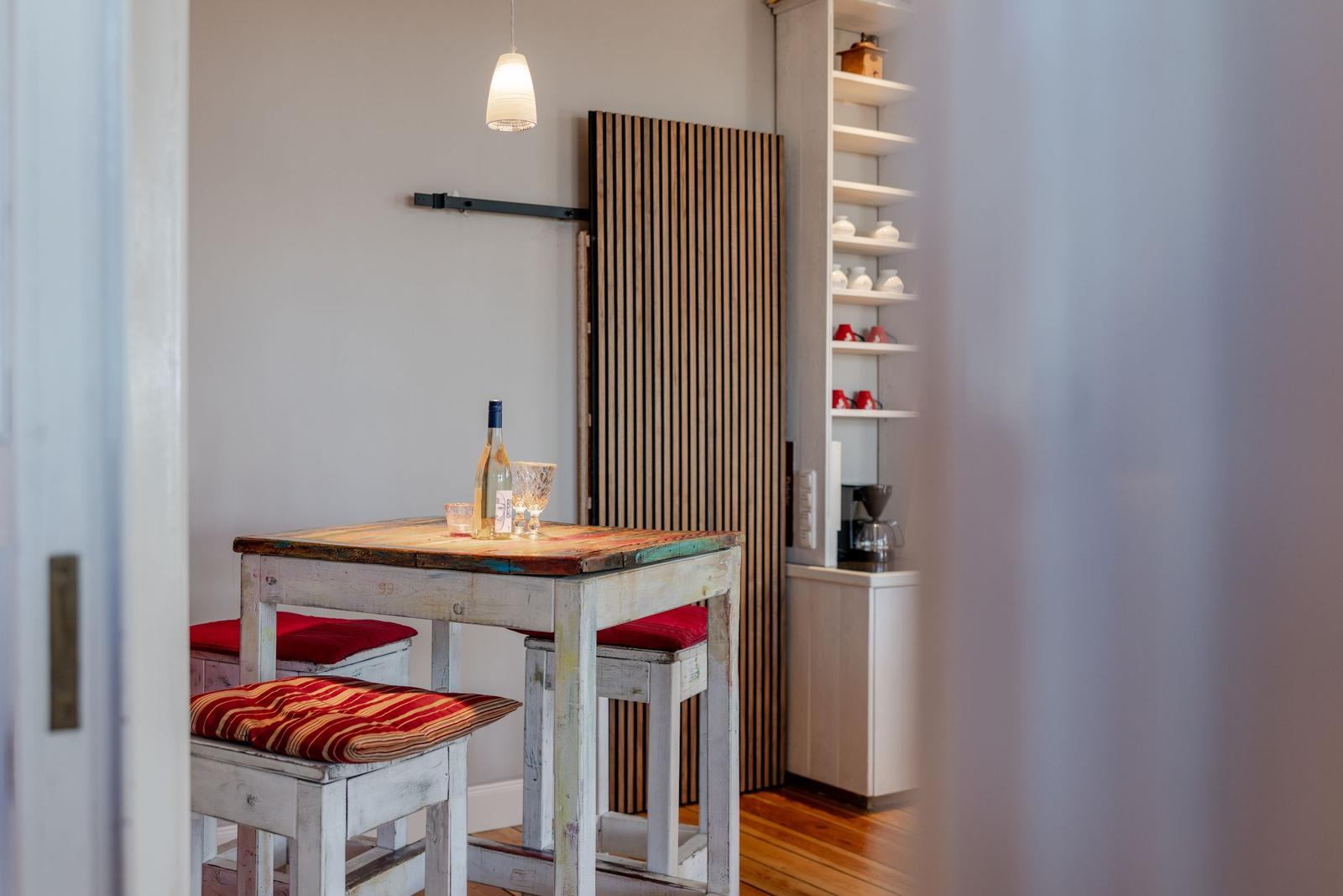 Esstisch mit Stühlen und einer Weinflasche. Hintergrund: Holzpaneel und Regal mit Geschirr.