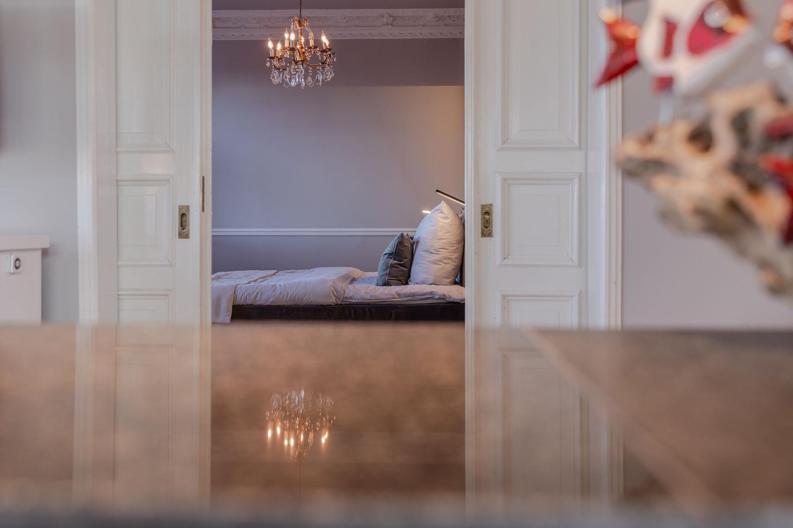Schlafzimmer mit Bett, Kissen und Kronleuchter. Durchgehendes Licht und hölzernes Bodenbelag.