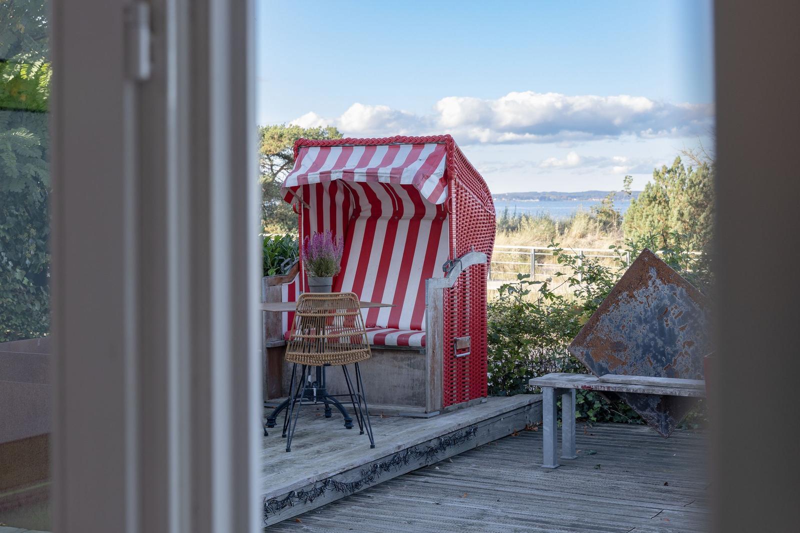 Terrasse mit Strandkorb, Tisch und Stuhl, Blick auf Wasser und Bäume.