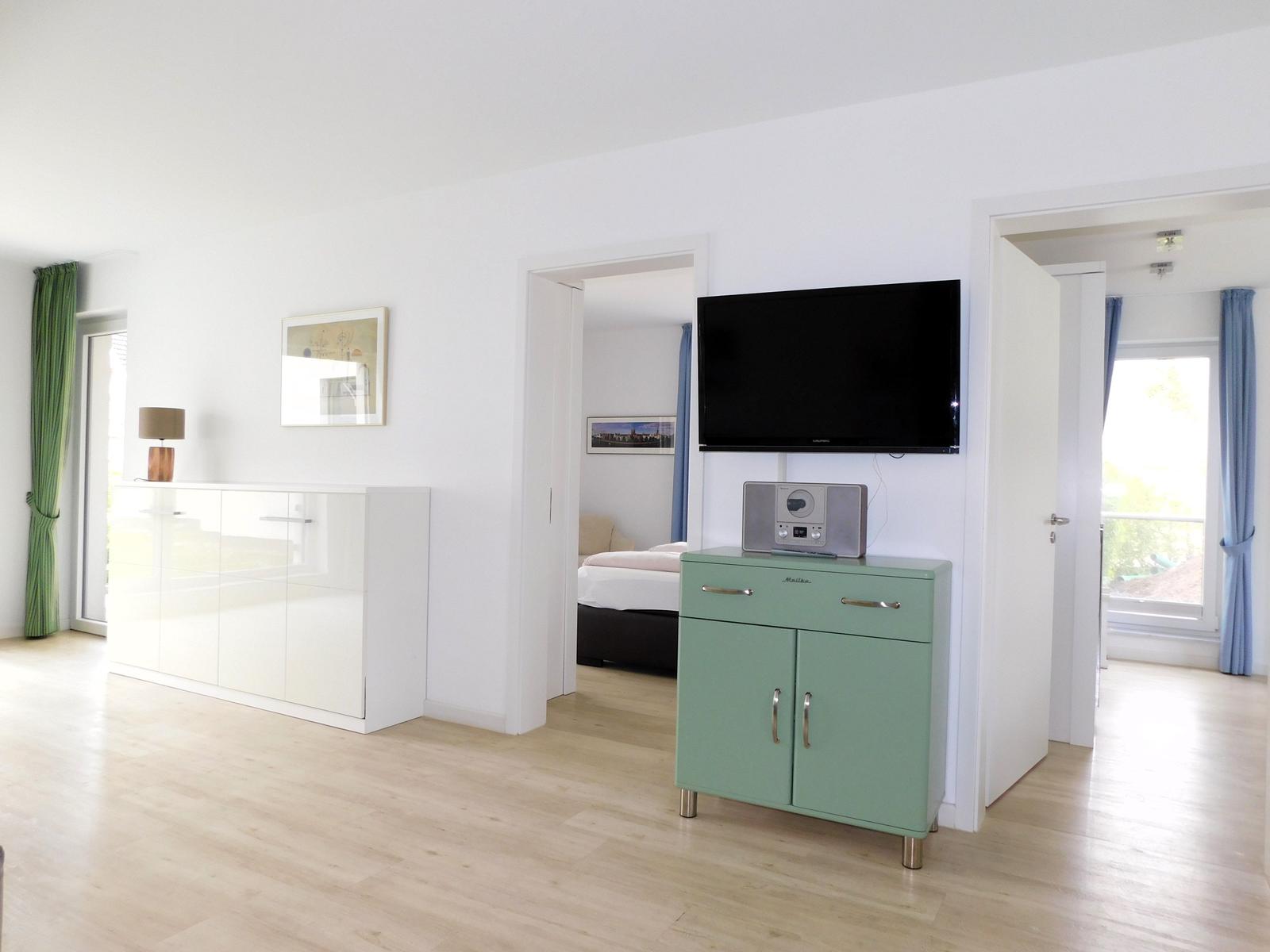 Wohnzimmer mit Fernseher und grünem Schrank. Tür führt in Schlafzimmer.