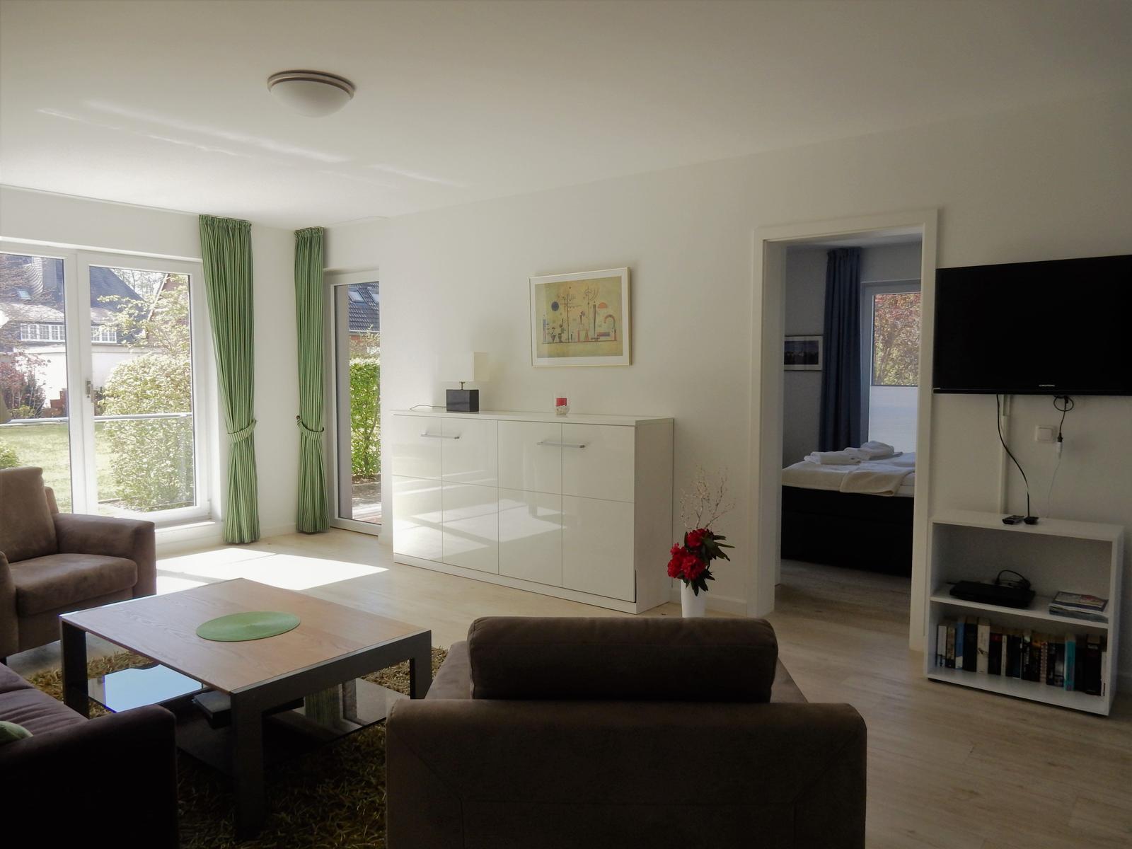 Wohnzimmer mit Sofa, Couchtisch und Blick auf Garten. Fernseher und Schlafzimmer sichtbar.