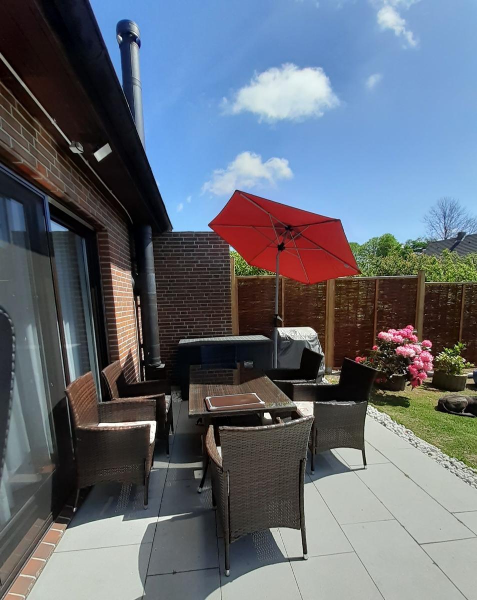 Terrasse mit Tisch, Stühlen und rotem Sonnenschirm. Grasfläche und Blumen im Hintergrund.